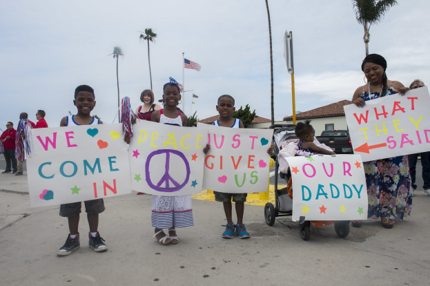 Gruppe von Kindern mit Schildern 'Wir kommen in Frieden, gebt uns unseren Daddy' auf einer Straße vor einem Gebäude, mit einer Frau, die einen Kinderwagen schiebt, und Bäumen im Hintergrund.