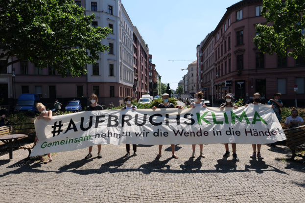 Eine Gruppe von Menschen mit Masken, die ein Transparent mit der Aufschrift "Aufbruchsklima" tragen, während sie an einer Klimaprotestaktion in Berlin, Deutschland, teilnehmen, umgeben von urbanen Elementen wie Bänken, Bäumen und Fahrzeugen.