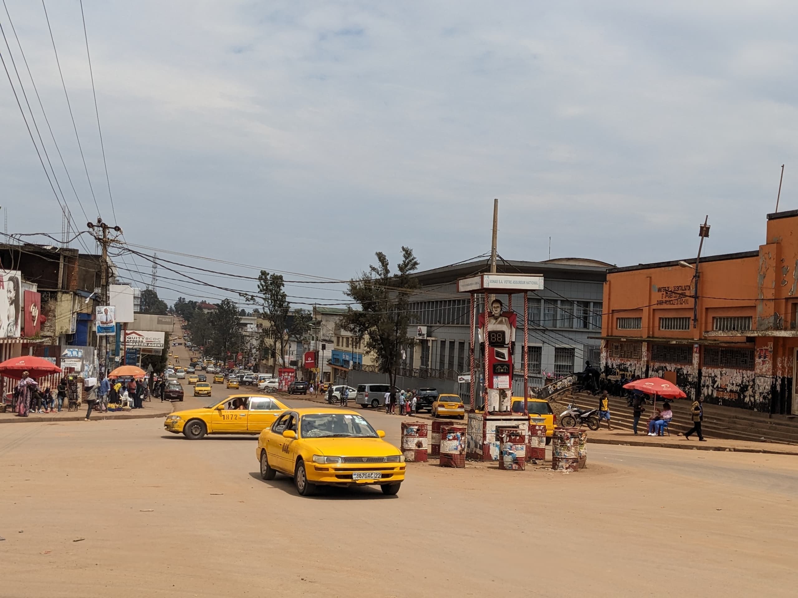 Ein gelbes Taxi fährt eine belebte Stadtstraße mit Gebäuden an den Seiten hinunter, während Fußgänger mit Schirmen auf dem Bürgersteig unter einem bewölkten Himmel gehen.