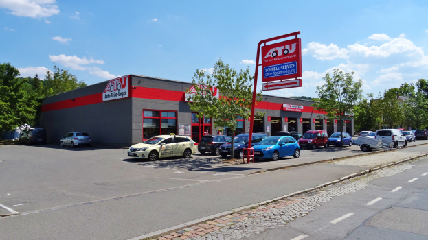 Autohaus mit parkenden Autos, umgeben von Gebäuden, Bäumen, Strommasten, Bauzäunen und einer Straße unter einem bewölkten Himmel.