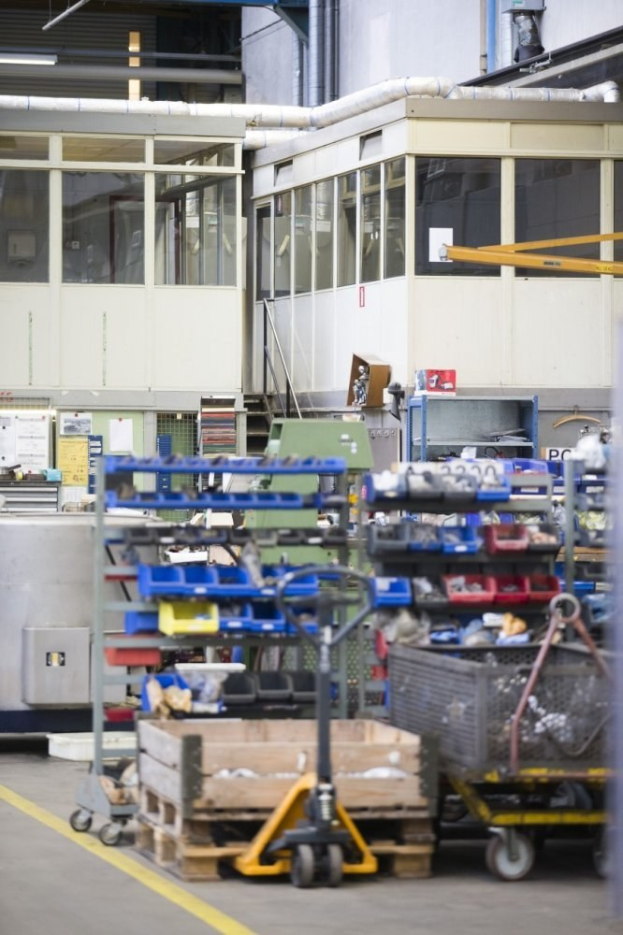 Ein großes Lager voller verschiedener Geräte wie Wagen, Schränke und eine Kiste, mit einer Wand aus Glasfenstern und Rohren im Hintergrund.