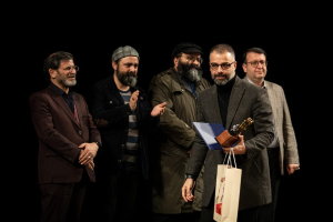 Eine Gruppe von Männern auf der Bühne der Eröffnungsnacht des Berliner Filmfestivals, einer hölt eine Tasche und eine Trophäe, mit einem dunklen Hintergrund.