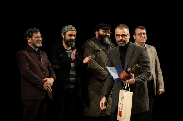 Eine Gruppe von Männern auf der Bühne der Eröffnungsnacht des Berliner Filmfestivals, einer hölt eine Tasche und eine Trophäe, mit einem dunklen Hintergrund.
