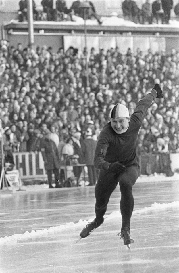 Altes Schwarz-Weiß-Foto eines Mannes, der auf einer Eisbahn Schlittschuh läuft und eine Mütze und Handschuhe trägt, mit unscharfen Figuren und Strukturen im Hintergrund.