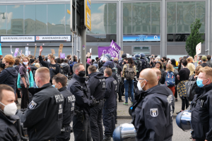 Große Menschenmenge vor einem Gebäude protestierend, einige halten Schilder und tragen Helme, mit einem Schildständer und einem Baum im Hintergrund.
