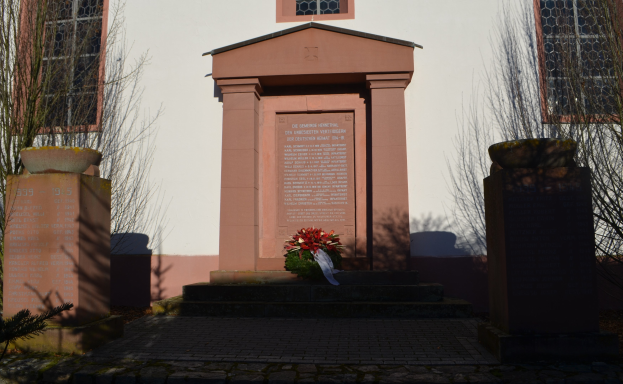 Ein Gebäude mit Fenstern, ein Denkmal mit Text, ein Blumenstrauß auf dem Boden, Topfpflanzen, eine Baumgruppe, ein bewölkter Himmel und ein Grab mit einem Kranz vor dem Gebäude.