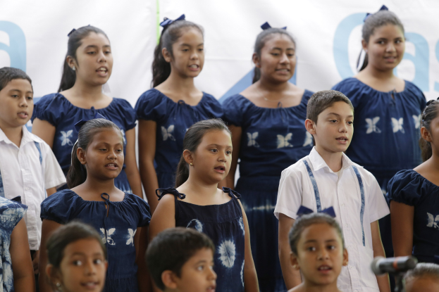 Gruppe von Kindern in einem Schulchor, der vor einem Mikrofon singt, mit einem Banner im Hintergrund.