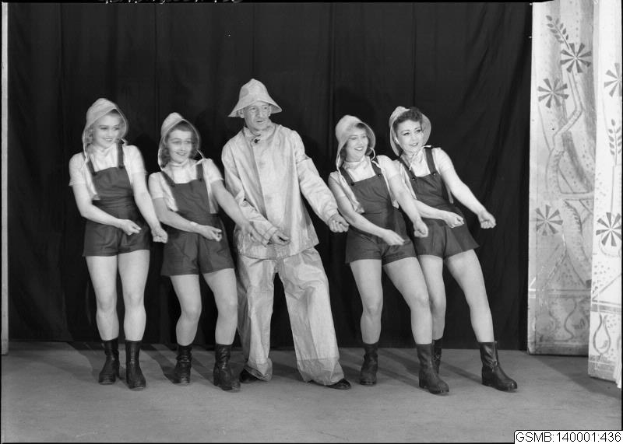 Schwarz-weiß-Fotografie von fünf Menschen in Hüten, die auf einer Bühne mit einem Vorhanghintergrund stehen, mit der Beschriftung "1940er-Jahre-Tanztruppe" unten.