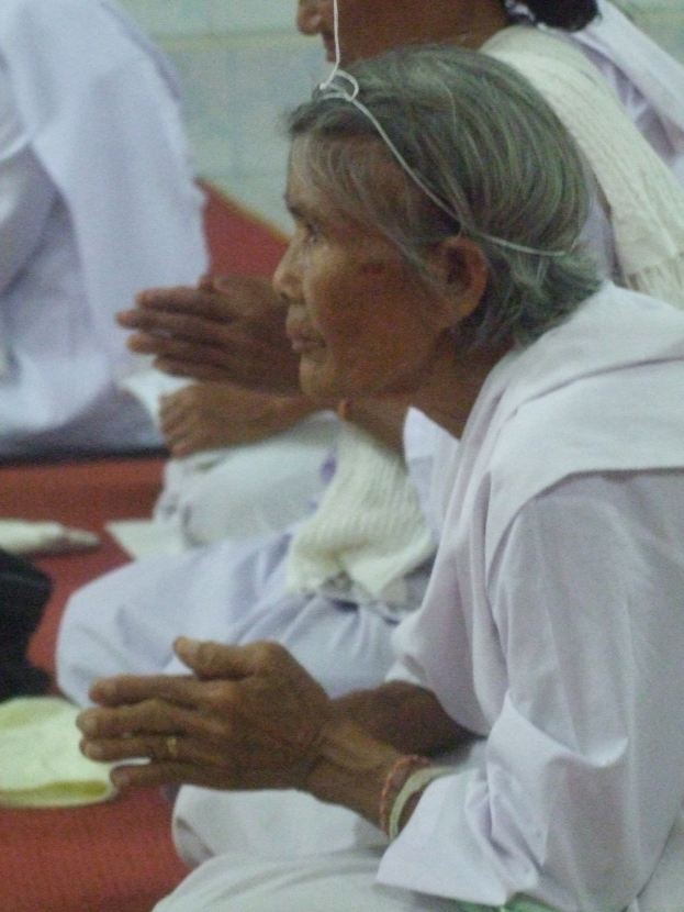 Alte Frau im Krankenhausbett liegend, Hände gefaltet, Augen geschlossen und Kopf auf den Händen ruhend, umgeben von Menschen im Hintergrund.