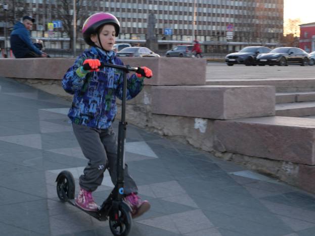 Ein junger Junge mit Helm und Handschuhen fährt auf einem Gehweg eine Treppe hinunter, mit Fahrzeugen, Menschen, Bäumen, Pfählen, Brettern, Gebäuden und einem klaren blauen Himmel im Hintergrund.