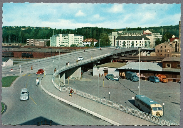 Altes Schwarz-Weiß-Foto einer Stadtstraße mit Autos, Bussen, Fußgängern auf einer Brücke, Laternen, mehrstöckigen Gebäuden mit Fenstern, Bäumen und einem bewölkten Himmel.
