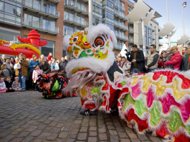 Ein lebendiges chinesisches Neujahrsfest in Amsterdam mit einem Löwen tanzen im Vordergrund und einer Menge von Menschen, einige halten Kameras, die sich darum versammelt haben, vor einem Hintergrund von Gebäuden, Straßenlaternen und einem klaren blauen Himmel.