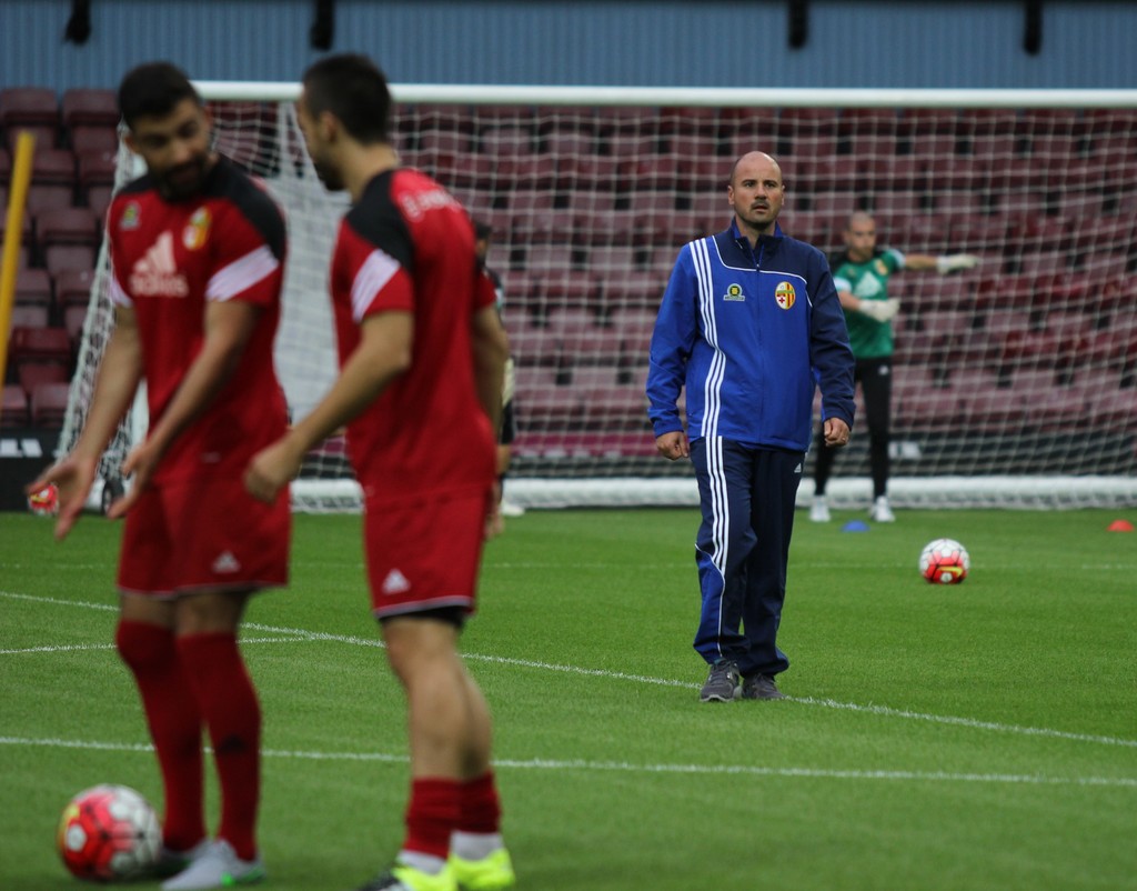 Ein Fußballfeld mit darauf stehenden Spielern, zwei Bällen und einer leeren Tribüne im Hintergrund.