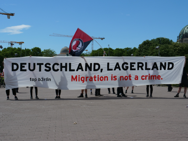 Eine Gruppe von Menschen, die auf dem Boden stehen und ein Banner halten, auf dem "Deutschland, Lagerland Migration ist kein Verbrechen" und eine Flagge zu sehen ist, mit Straßenlaternen, Bäumen, Kränen, einem Gebäude und einem bewölkten Himmel im Hintergrund.