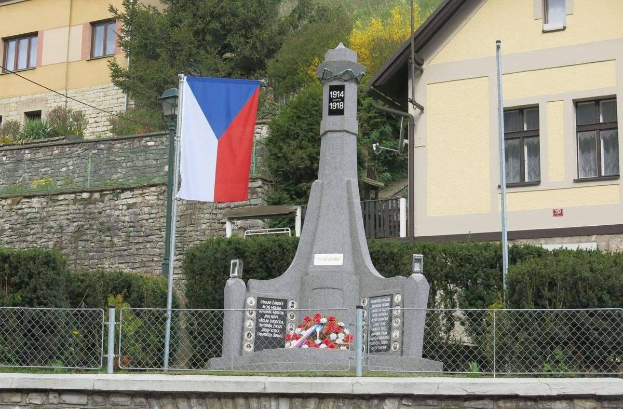 Ein Denkmal mit einer Fahne darauf, umgeben von einem Zaun, Pflanzen, Bäumen und einer Wand, dient als Gedenkstätte für die Opfer des Zweiten Weltkriegs, mit einer Inschrift darauf.