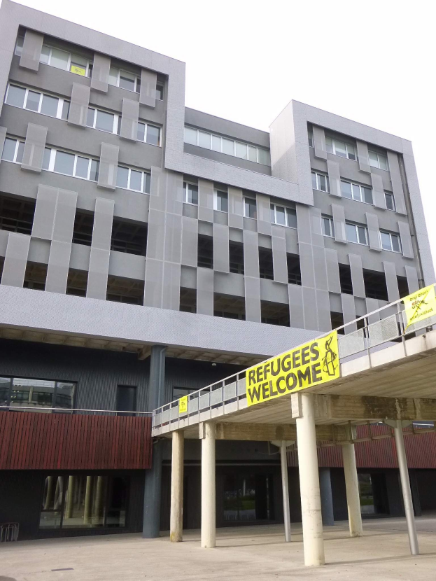 Großes Gebäude mit einem "Geflüchtete Willkommen"-Schild, Säulen, Geländer und Himmel im Hintergrund.