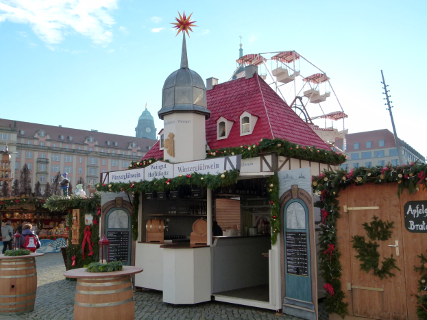 Ein kleines Gebäude mit einem roten Dach und einem Riesenrad davor, umgeben von Festdekorationen und Menschen auf einem Oktoberfest in München, Deutschland.