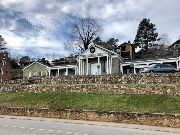 Steinwand mit parkenden Autos, benachbarten Gebäuden mit Fenstern und Türen, Bäumen und einem bewölkten Himmel im Hintergrund, als Weltkulturerbe-Museum bezeichnet.