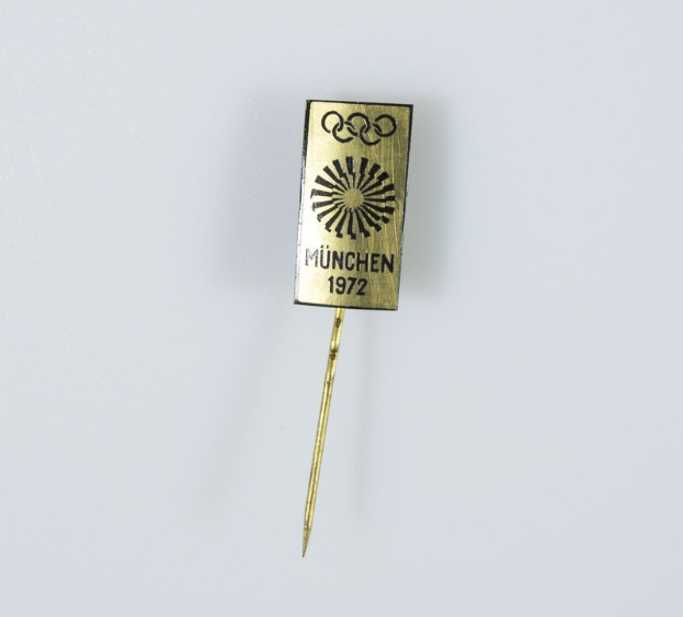 Goldene Nadel mit 'München 1972 Olympische Spiele' Aufschrift auf weißem Hintergrund.