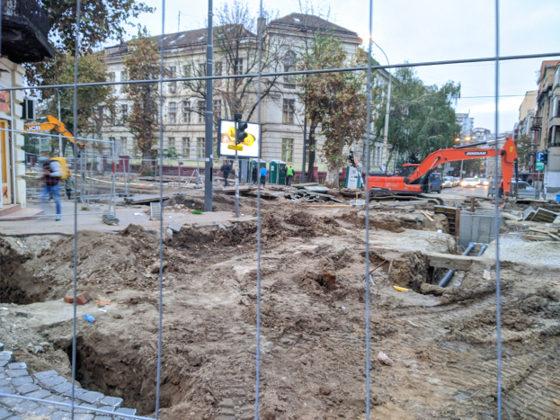 Baustelle mit einem ausgehobenen Bereich, der von einem Metallzaun umgeben ist, angrenzende Straße mit Fahrzeugen, Fußgänger auf einem Gehweg, Verkehrsampeln, Schilder, Bäume, Gebäude mit Fenstern und ein klarer blauer Himmel.