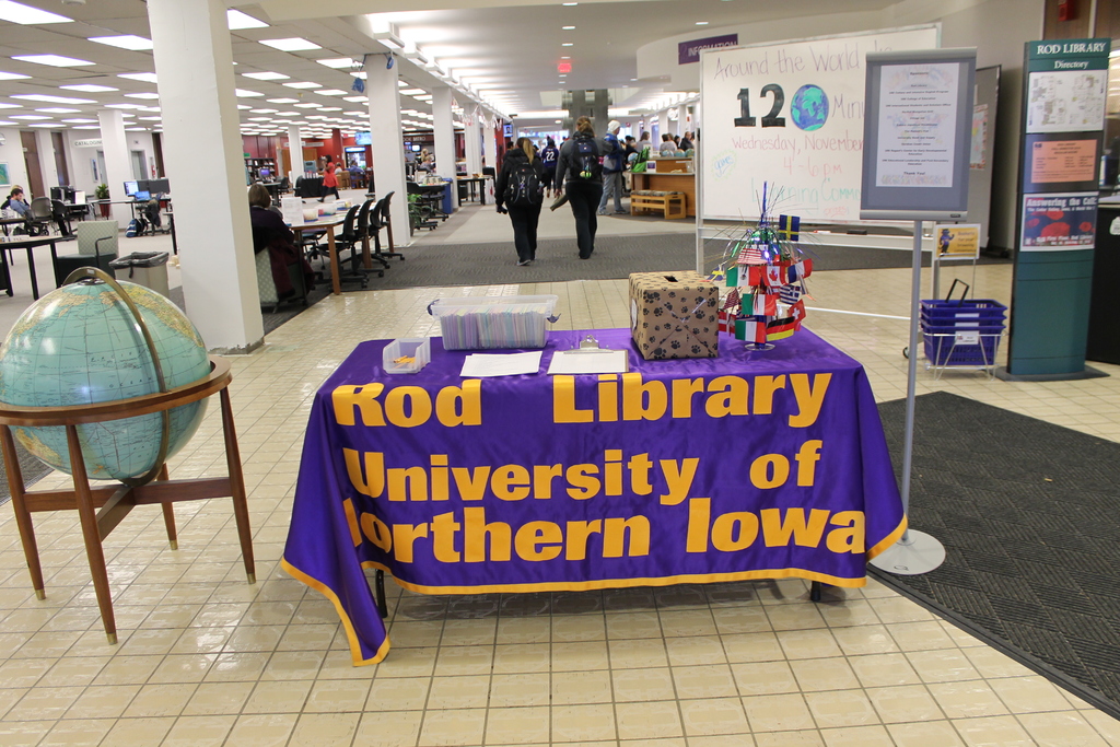 Eine Ausstellung mit einem Tisch, auf dem verschiedene Landesflaggen, eine Box, Papiere und ein Banner liegen, eine Kugel links daneben, ein Anschlagbrett rechts daneben und mehrere Tische und Stühle im Hintergrund.