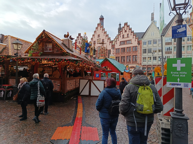 Eine Gruppe von Menschen geht eine Kopfsteinpflasterstraße neben einem Weihnachtsmarkt in Nürnberg, Deutschland, entlang, wobei einige Taschen, Laternenmasten und Schilder mit Text tragen, während Gebäude mit Fenstern und ein bewölkter Himmel im Hintergrund zu sehen sind.