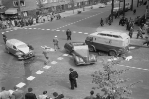 Eine Schwarz-Weiß-Szene eines Autounfalls an einer Straßecke mit mehreren Fahrzeugen, darunter ein Van, einer Gruppe von Menschen drumherum und Gebäude, Bäume, Masten und Texttafeln im Hintergrund.