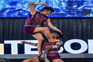 Ein Mann und eine Frau in violetten Kleidern führen Akrobatik auf der Bühne aus, wobei der Mann die Frau in seinen Armen hält, während das Publikum unten zusieht und ein schwarzer Vorhang und ein Bildschirm im Hintergrund zu sehen sind.