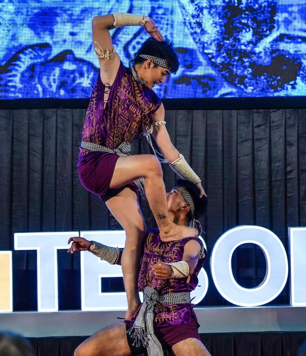 Ein Mann und eine Frau in violetten Kleidern führen Akrobatik auf der Bühne aus, wobei der Mann die Frau in seinen Armen hält, während das Publikum unten zusieht und ein schwarzer Vorhang und ein Bildschirm im Hintergrund zu sehen sind.