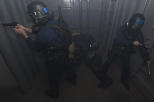 Eine Gruppe von Polizisten in Einsatzausrüstung, die zusammenstehen, einer hält eine Waffe, mit einem Metallcontainer und einer Leiter im Hintergrund.