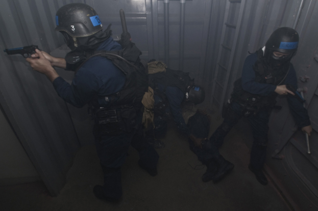 Eine Gruppe von Polizisten in Einsatzausrüstung, die zusammenstehen, einer hält eine Waffe, mit einem Metallcontainer und einer Leiter im Hintergrund.