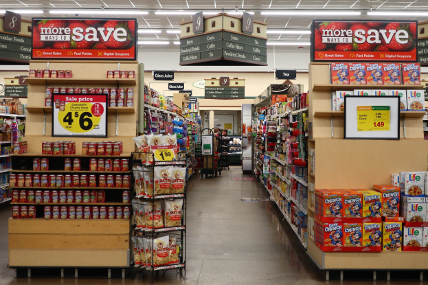 Ein Kiosk-Innenraum mit verschiedenen Lebensmittelartikeln, die auf Gestellen und dem Boden angeordnet sind, beleuchtet von Deckenlampen und mit Informationsschildern mit Text und Zahlen.