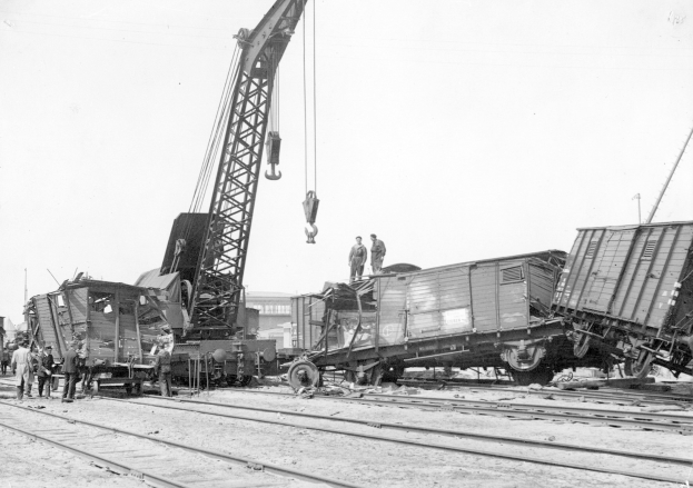 Schwarzes und weißes Bild einer Zugkatastrophe mit einem Kran, der einen Zugwagen auf die Schienen hebt, umgeben von Menschen, Gebäuden und einem klaren Himmel.