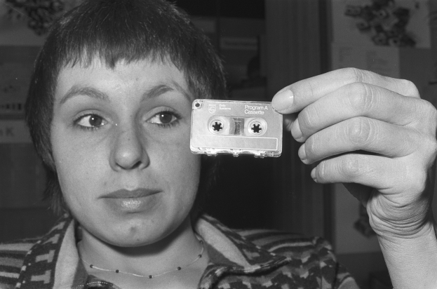 Eine Frau hält eine Kassette vor ihr Gesicht an einer Wand, dargestellt in Schwarz-Weiß.