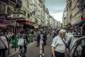 Zwei große Gebäude auf beiden Seiten einer Straße mit einer Menge Menschen dazwischen, Stände im Erdgeschoss, einige Mülltonnen, ein Mann auf einem Fahrrad und geparkte Autos auf der gegenüberliegenden Seite.