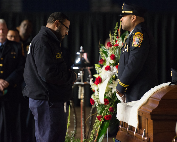 Zwei Polizisten in Uniform stehen feierlich neben einem mit Blumen geschmückten Sarg, mit wenigen Personen im Hintergrund.