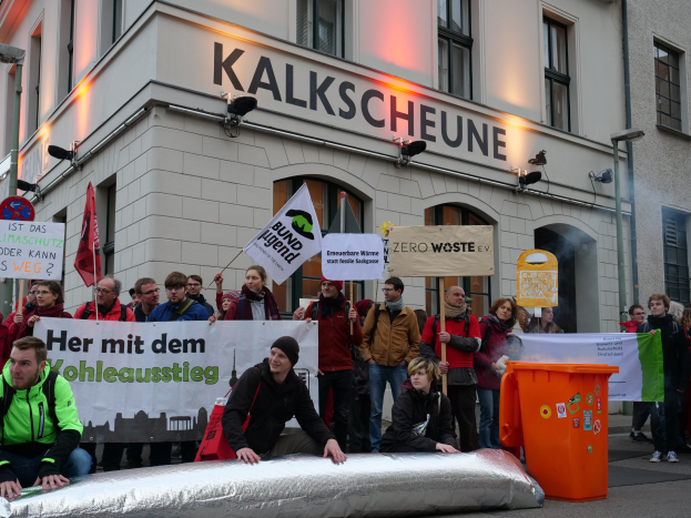 Eine Gruppe von Menschen mit Schildern und Plakaten vor einem Gebäude während einer Demonstration in Deutschland, mit zwei Personen im Vordergrund und einem Mücllimer auf der rechten Seite.