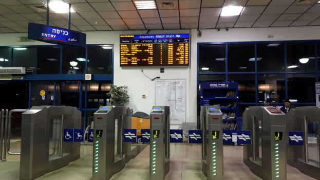 Ein Mann steht vor einer Metalldrehkreuz an einem Flughafen, umgeben von Metalldetektoren, Informationsschildern, einem Topf mit einer Pflanze, einer Wandtafel, einer Decke mit Leuchten und einem Anzeigebildschirm.