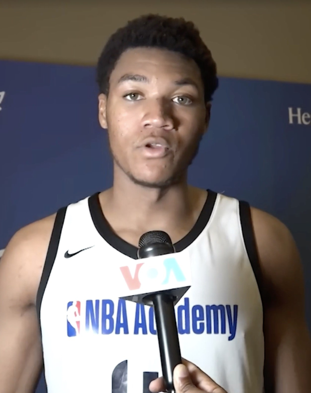 Ein Mann in einem weißen Tanktop spricht bei einer Medienveranstaltung zum NBA All-Star Game 2019 in ein Mikrofon, mit einem Banner und einer Wand im Hintergrund.