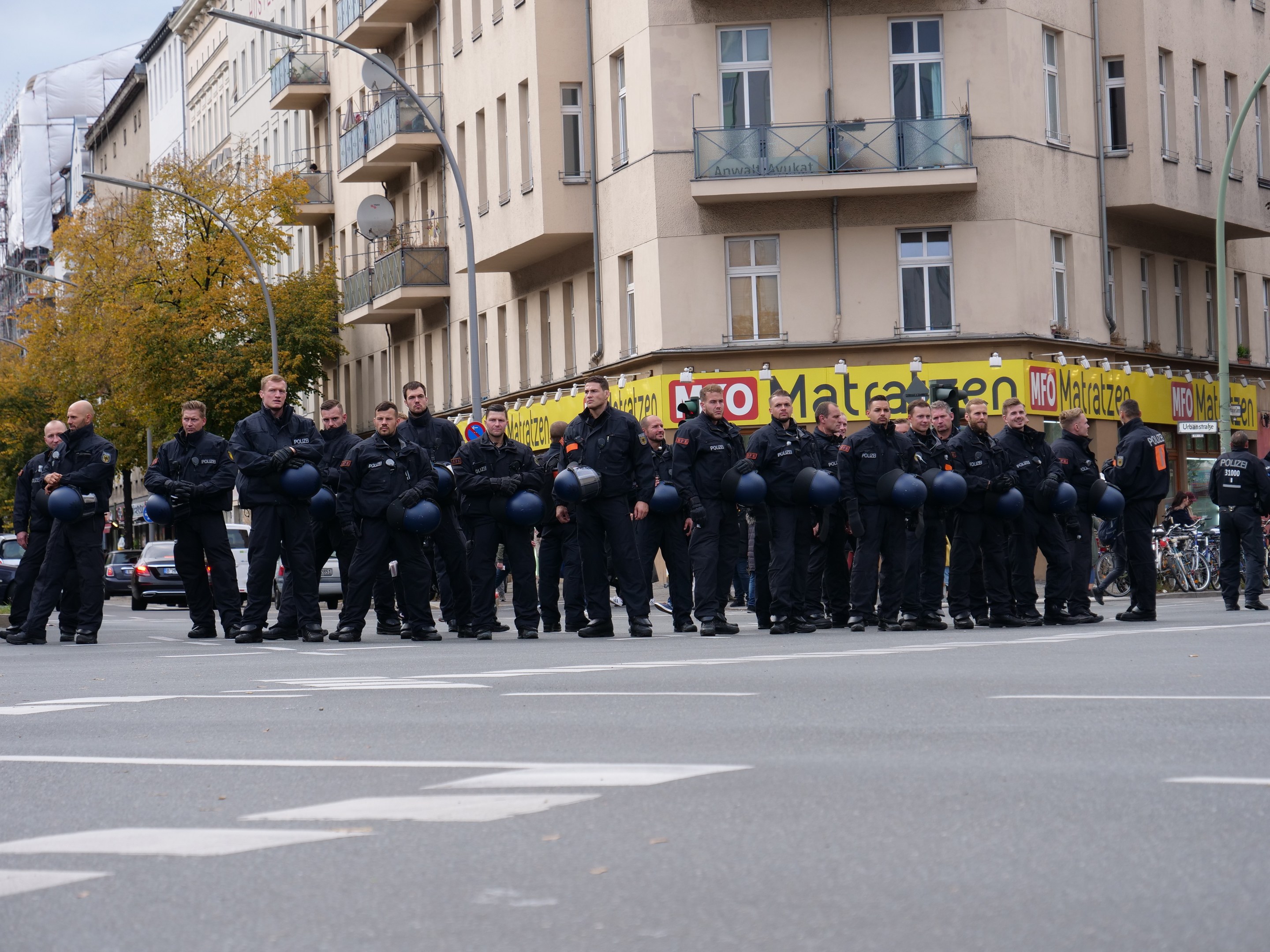 Eine Gruppe von Polizisten in schwarzen Uniformen mit blauen Helmen steht in einer Straße mit Laternen, Bäumen und glasfensterigen Gebäuden unter einem klaren blauen Himmel.