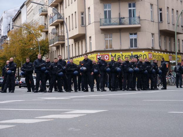 Eine Gruppe von Polizisten in schwarzen Uniformen mit blauen Helmen steht in einer Straße mit Laternen, Bäumen und glasfensterigen Gebäuden unter einem klaren blauen Himmel.