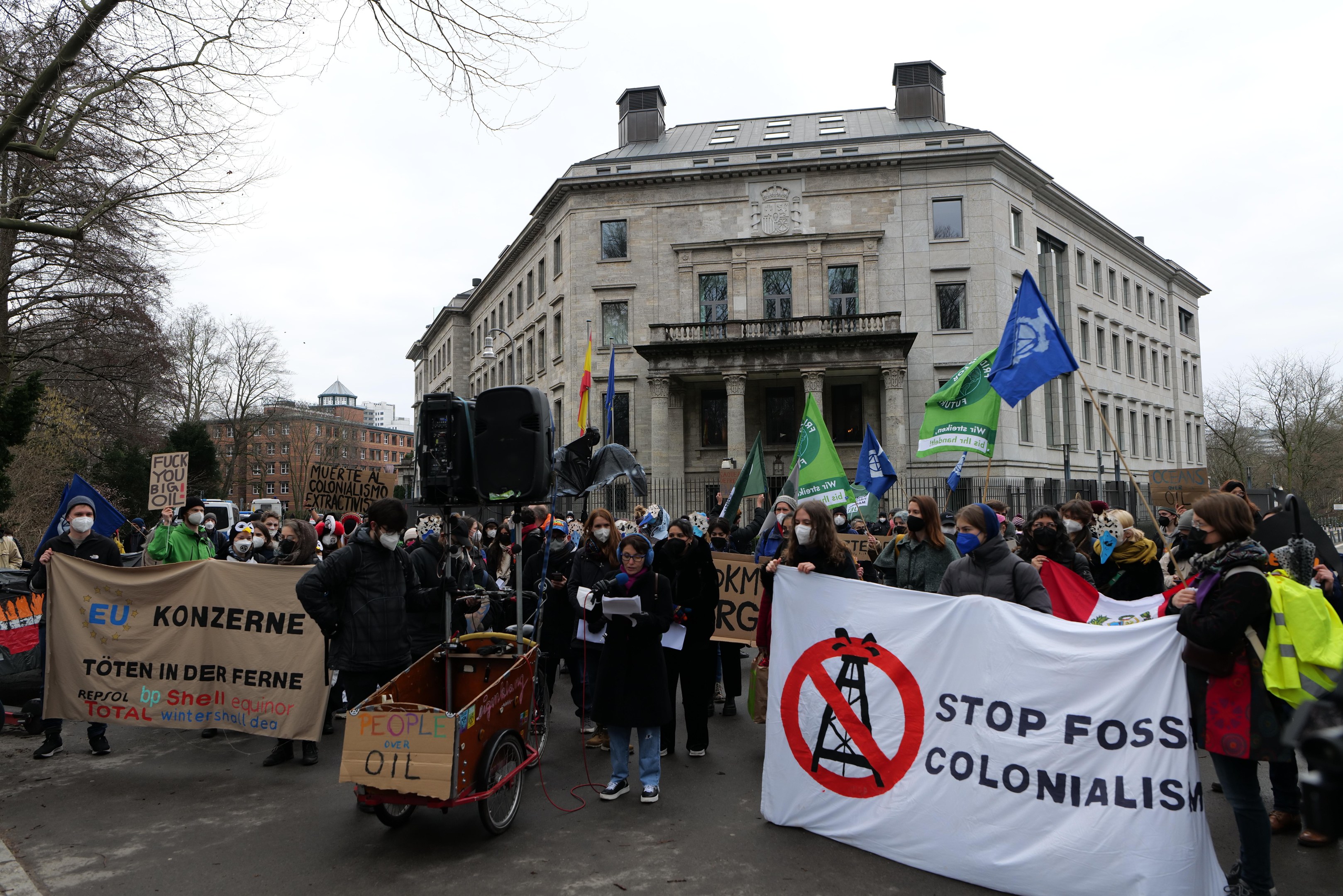 Eine große Gruppe von Menschen marschiert mit Transparenten und Fahnen eine Straße entlang, vorne ein Fahrzeug, protestierend gegen fossile Brennstoffe; Bäume, Gebäude mit Fenstern und ein klarer blauer Himmel sind im Hintergrund zu sehen.
