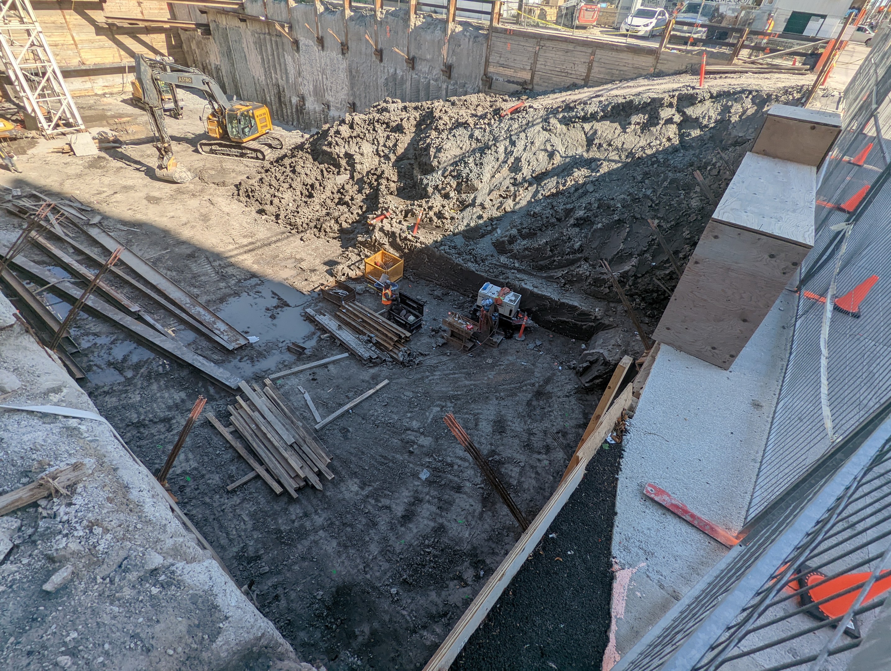 Baustelle mit einem großen Haufen Erde, umgeben von Kränen, Holzstangen und anderem Equipment, begrenzt von einem Maschendrahtzaun, mit Gebäuden, Fahrzeugen, Pfählen, Bäumen und Himmel im Hintergrund.