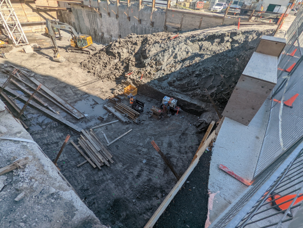 Baustelle mit einem großen Haufen Erde, umgeben von Kränen, Holzstangen und anderem Equipment, begrenzt von einem Maschendrahtzaun, mit Gebäuden, Fahrzeugen, Pfählen, Bäumen und Himmel im Hintergrund.