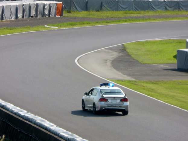 Ein Streifenwagen fährt auf einer Rennstrecke mit Gras an beiden Seiten, mit Menschen, Zelten und anderen Gegenständen im Hintergrund.