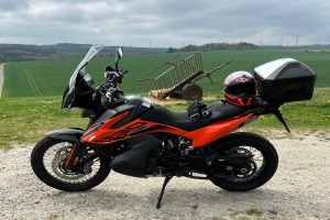 Ein KTM 1290 Super Adventure R Motorrad auf einem Schotterweg mit einem Helm und einem Gepäckwagen daneben, umgeben von grünem Gras, hohen Bäumen und Strommästen unter einem bewölkten Himmel.