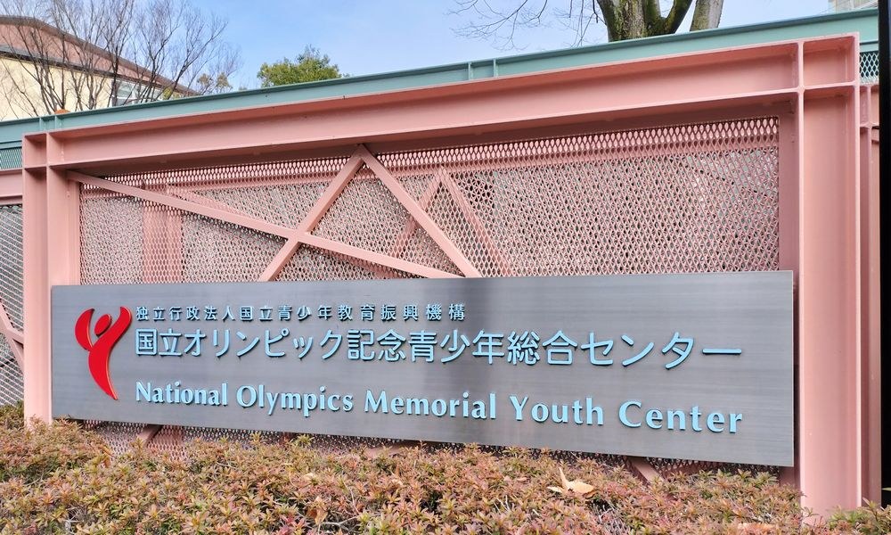 Das Nationale Olympische Memorial Youth Center mit einer Wandtafel, Pflanzen am Boden, Bäumen und Gebäuden im Hintergrund und einem klaren blauen Himmel darüber.