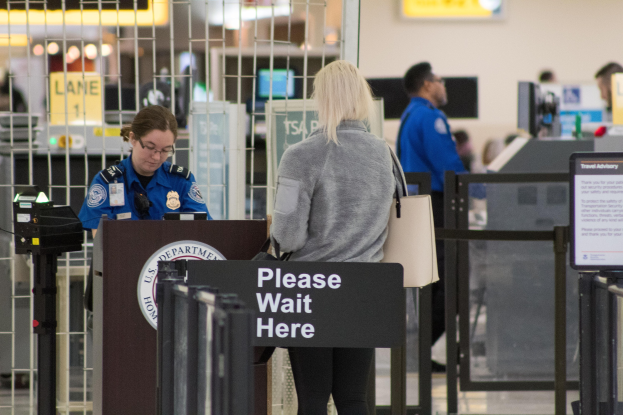 Eine Frau in einer grauen Jacke und schwarzen Hosen steht an einem Flughafen-Metallsensor und hält eine Handtasche, neben ihr steht ein Polizeibeamter in einer blauen Uniform und hinter ihr eine Tafel.
