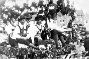 Eine Gruppe von Menschen in traditioneller bayrischer Tracht fährt auf einem mit Laub geschmückten Festwagen in einem Umzug mit, einige halten Musikinstrumente, mit Gebäuden und einem klaren Himmel im Hintergrund, dargestellt in Schwarz-Weiß.