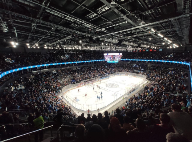 Eishockeyspiel in einer großen Arena mit Zuschauern in den Rängen und Personal am Boden, beleuchtet von Scheinwerfern, mit einem Bildschirm oben.